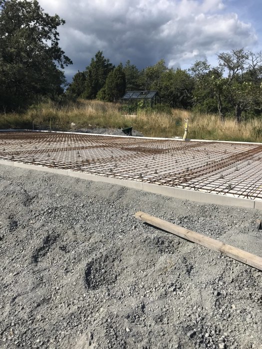 Grundläggning med armeringsjärn, skottkärra, skogsmark, växthus och mulet himmel i bakgrunden.