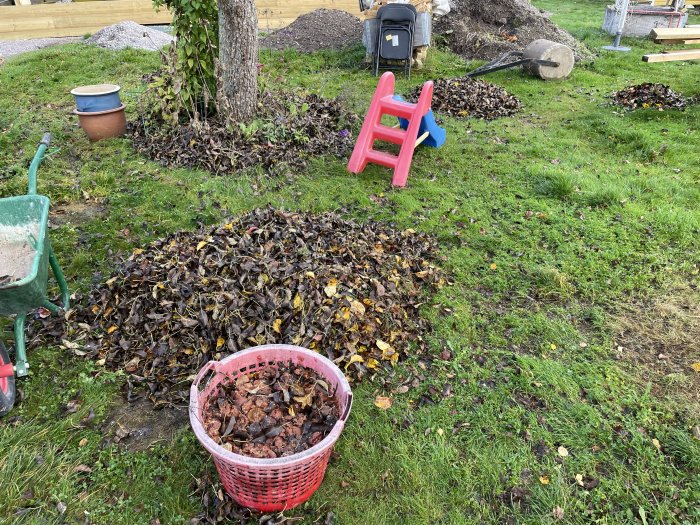 Trädgård med lövhögar, skottkärra, barnstol, leksak, krukor och gräsmatta i dagsljus.