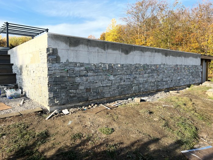 En grå betongmur under konstruktion med stenklädnad, byggmaterial runt, mot höstträd bakgrund.