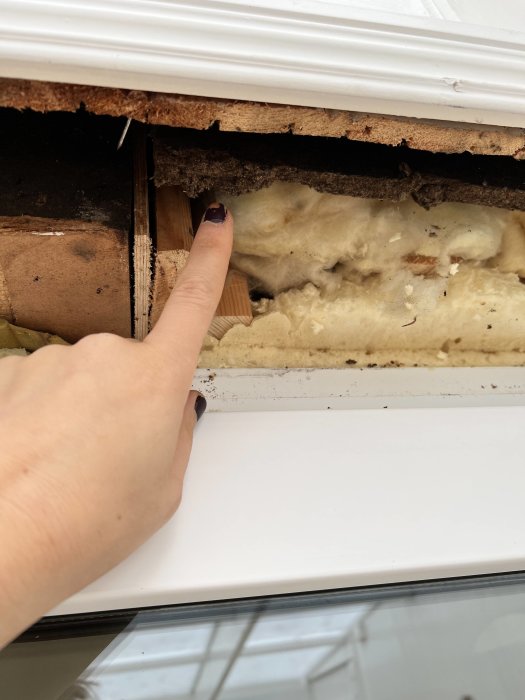 En hand pekar på isolering i en skadad fönsterkarm med synliga träbitar och isoleringsmaterial.