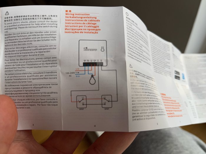 En hand håller en manual med elektrisk installationsinstruktion på olika språk, med en schematisk diagram.