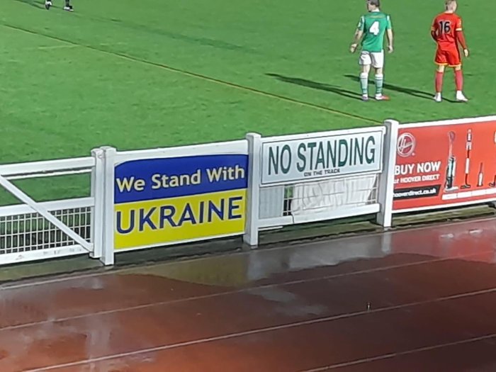 Fotbollsplan med ironisk kombination av "We Stand With UKRAINE" och "NO STANDING" skyltar, två spelare i bakgrunden.