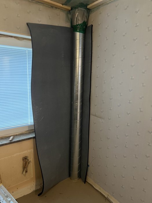 Luftkanal genom fönster, ventilationsslang, blå persienn, tapetvägg, delvis renoverat rum, matta saknas.