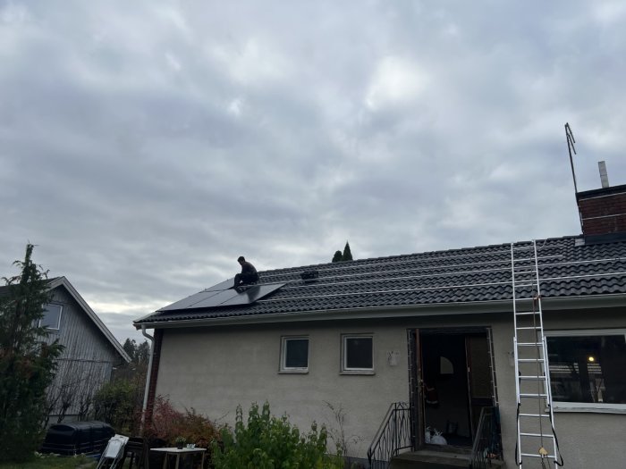 En person på ett hustak installerar solpaneler, molnig himmel, stege, bostadshus, trädgård med vegetation och grill.