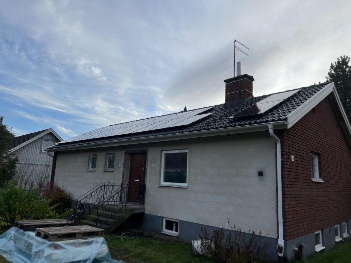 Enfamiljshus med röda tegel och vit puts, solpaneler på taket, byggmaterial framför, överväxande vegetation, grå himmel.
