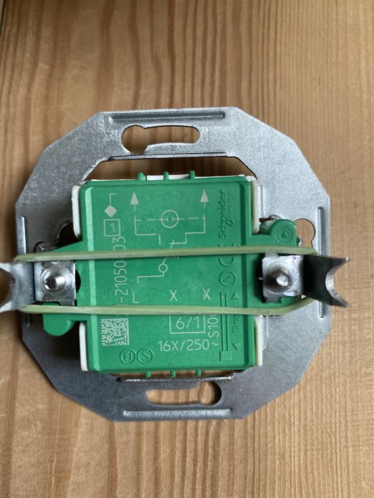 Grön och silverfärgad elektrisk komponent, troligen en strömbrytare eller vägguttag, med montagefästen och elektrisk information.