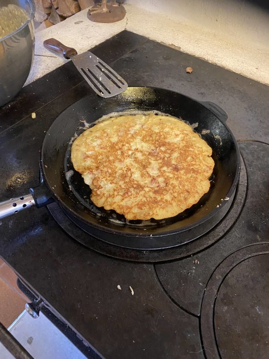 Stekt pannkaka i gjutjärnspanna på spishäll, med stekspade bredvid. Hyfsat gyllenbrun, hemlagad.