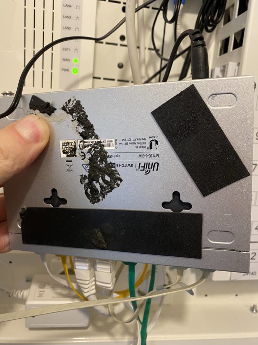 En hand håller en elektronisk apparat med slitna etiketter framför en nätverksutrustningspanel och kablar.