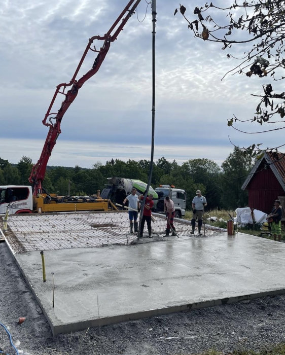 Betongplatta gjuts med betongpump; arbetare jämna ut och avlägsnar luftbubblor, byggarbetsplats, träd och molnig himmel.