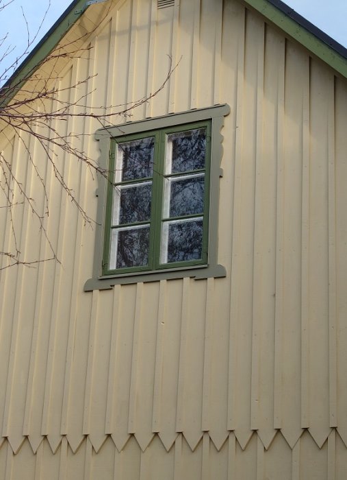 Gult trähus med vågformad panel, grönt fönster, reflektion av träd, blå himmel, del av gren framför.