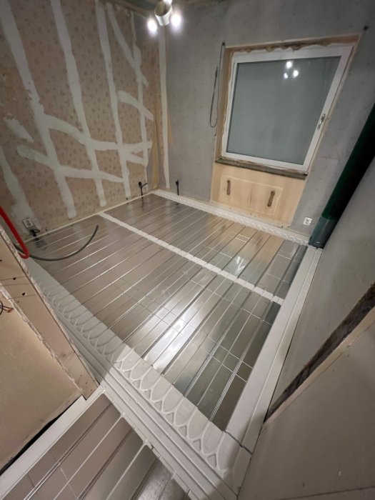 Ett rum under renovering med isolering synlig, fönster, ett värmegolv som installeras.