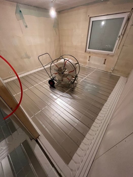 Ett outrett rum med fläkt, kabel, kala väggar, fönster och vit kaklad golv.