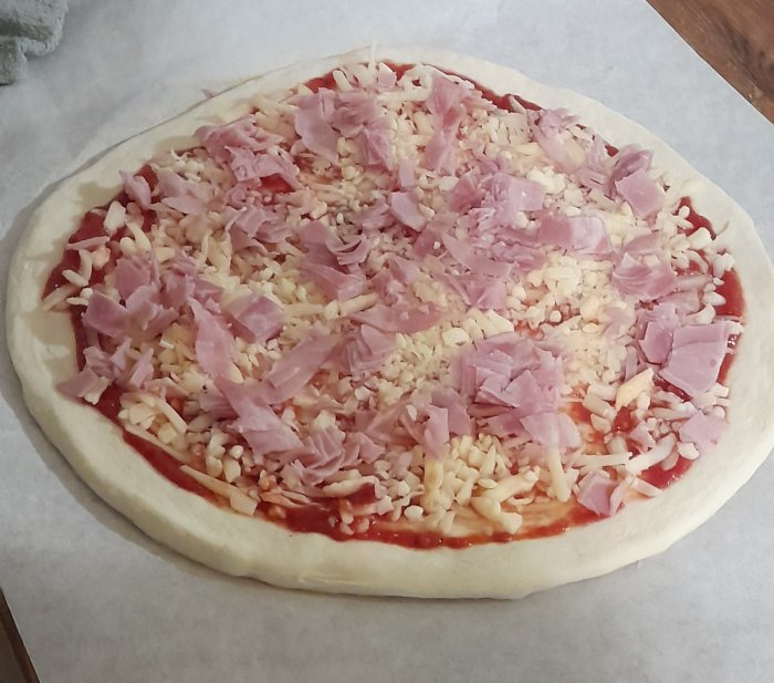 Ogräddad pizza med tomatsås, ost och skinka på bakplåtspapper.