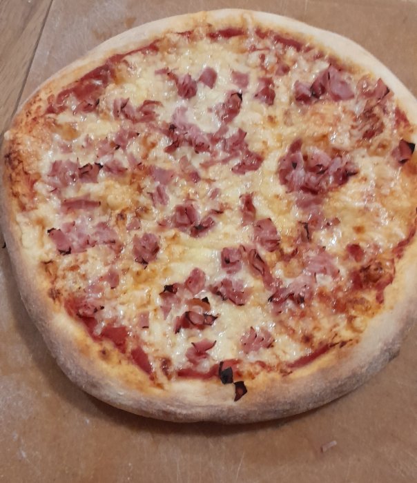 En bakad pizza med skinka och ost på ett brunt underlag.