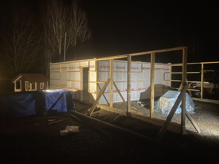 Byggarbetsplats på natten, oisolerad träkonstruktion, kallt ljus, stabiliserande stöttor, materialskydd under presenningar.