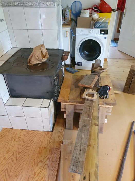 Renovering pågår i kök med gammal spis, tvättmaskin och verktyg utspridda runt träplankor och skyddspapp.