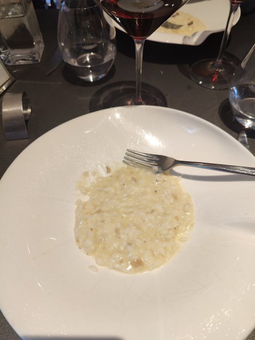 En portion risotto på en vit tallrik med gaffel, omgiven av vin och vattenglas.
