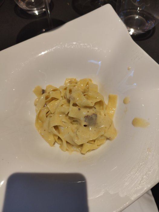 En portion pasta med sås på en vit tallrik, en vin- eller vattenglas synlig i bakgrunden.