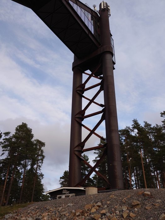 Utsiktstorn i metall med spiraltrappa, omgivet av tallskog och stenig mark. Skymning med moln på himlen.