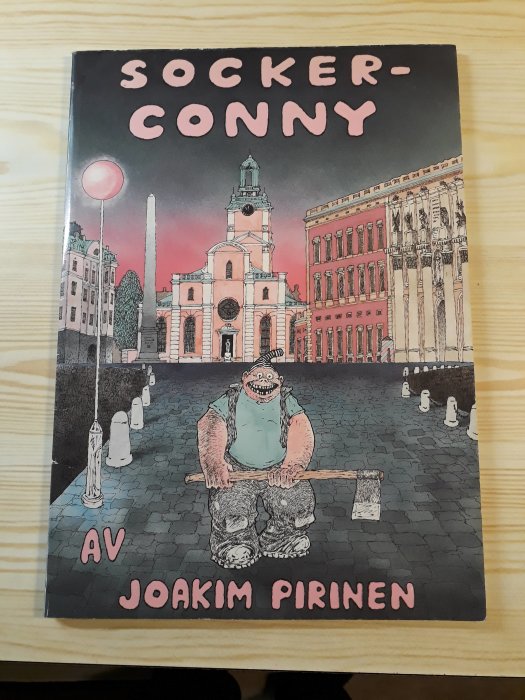 Omslag till "Socker-Conny" av Joakim Pirinen på ett bord, illustrerad karaktär framför klassisk arkitektur, humoristisk ton.