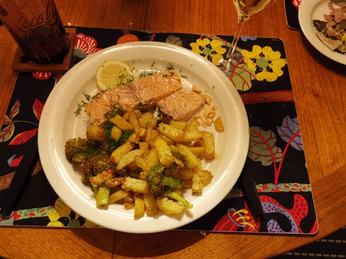 En tallrik med lax, potatis, broccoli och dill. Colaglas och ett glas vin. Mönstrad duk.