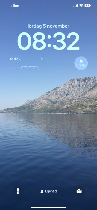 Skärmbild av en mobiltelefon visar klockan, datum, batteristatus över ett bergslandskap och havet.