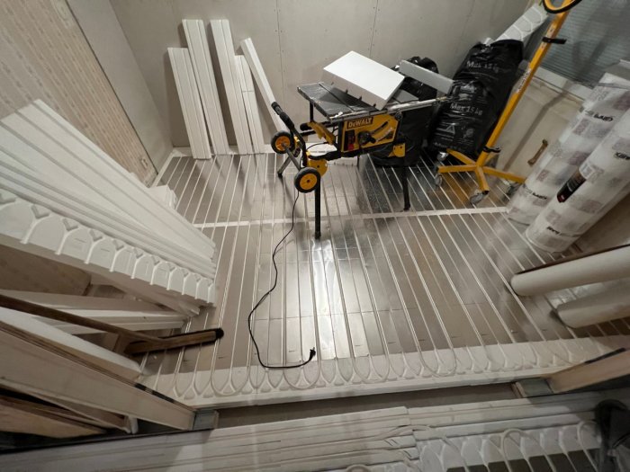 Byggplats inomhus med lister, isoleringsmaterial, verktyg och en såg på stativ. Pågående renovering eller byggnadsarbete.