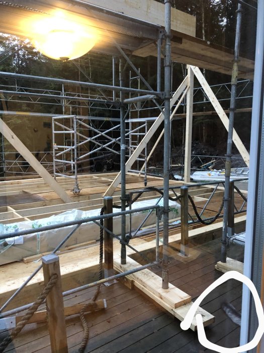 Byggarbetsplats vid skymning med byggnadsställningar, träreglar och tillfällig belysning, sedd genom ett fönster.