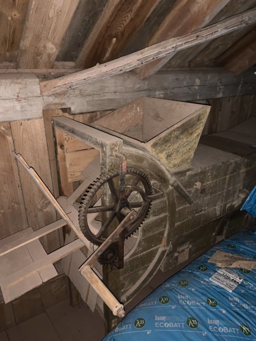 Gammal vind med kugghjulsmaskineri, träbjälkar, och isolering. Verkar vara ett loft eller vindrum.