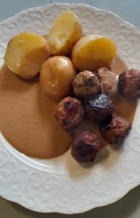 Köttbullar, potatis och brun sås på ett vitt, mönstrat tallrik.