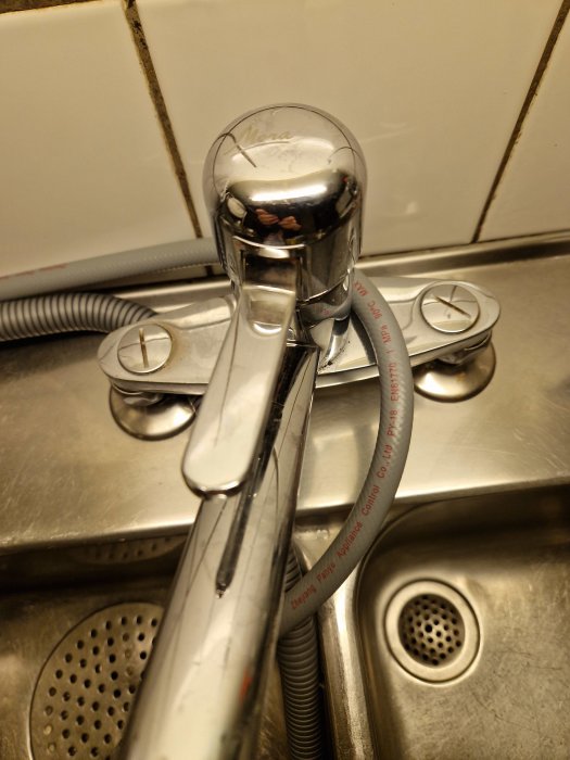 Kromad köksblandare, sladdar, diskho, kakelvägg, skruvar, märkning på slang, återspegling, lite smutsigt, rostfritt stål.
