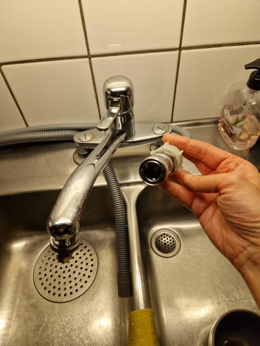 En hand håller en demonterad del av en köksblandare över ett rostfritt ståldiskho. Fliser bakgrund och diskmedel syns.