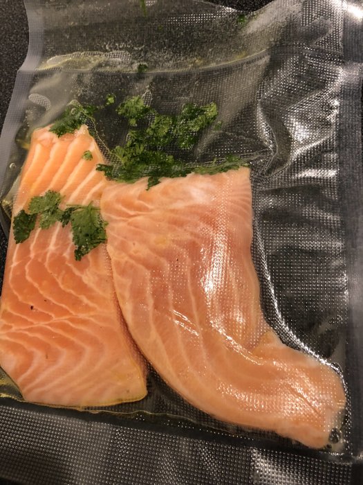 Två laxfiléer med färsk koriander, förpackade i vakuum, redo för tillagning eller förvaring.