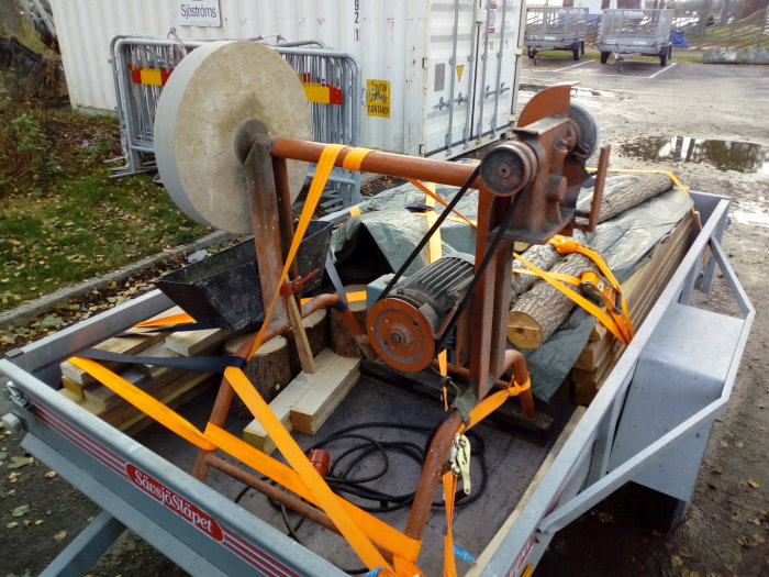 Ett lastat släp med virke och stenskärare, säkrat med orange spännband, utomhus vid container och fordon.