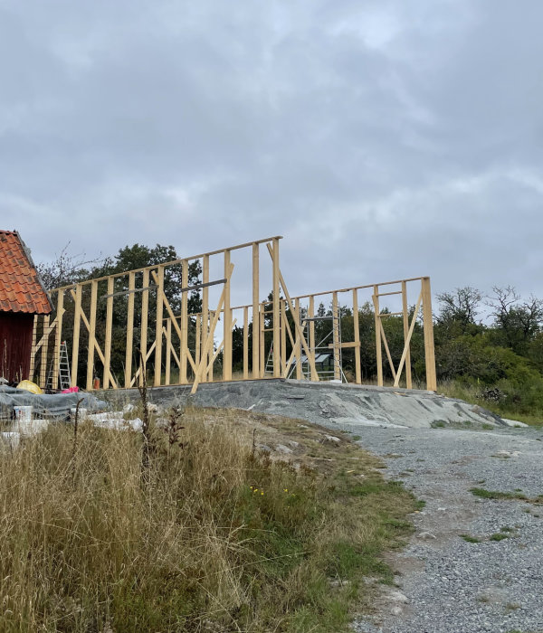 Trästomme för byggnad under konstruktion, klippgrund, molnig himmel, röd stuga delvis synlig, gräs och vild natur.