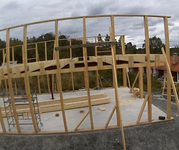 Träramkonstruktion av byggnad, pågående arbete, molnig himmel, röd byggnad i bakgrunden, grusig mark.