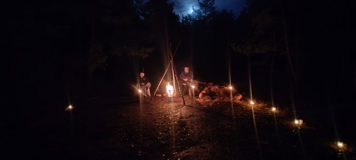 Två personer vid lägereld i skogen, månsken, små lyktor, mystisk och fredlig stämning.