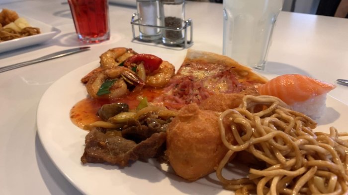 En mångsidig tallrik med blandad mat såsom pizza, sushi, räkor och nudlar. Inomhusmiljö.