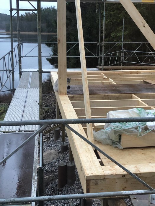 Byggarbetsplats med trästomme, ställning vid vatten, molnig himmel, pågående konstruktion, skog i bakgrunden.