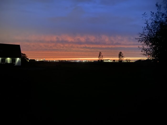 Solnedgång med orange himmel, konturer av hus och träd, ljus från fjärran stad, dimmig horisont.