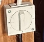 En vit, väggmonterad elektrisk strömbrytare med rund vridknapp, omgiven av fyra skruvar mot en träbakgrund.