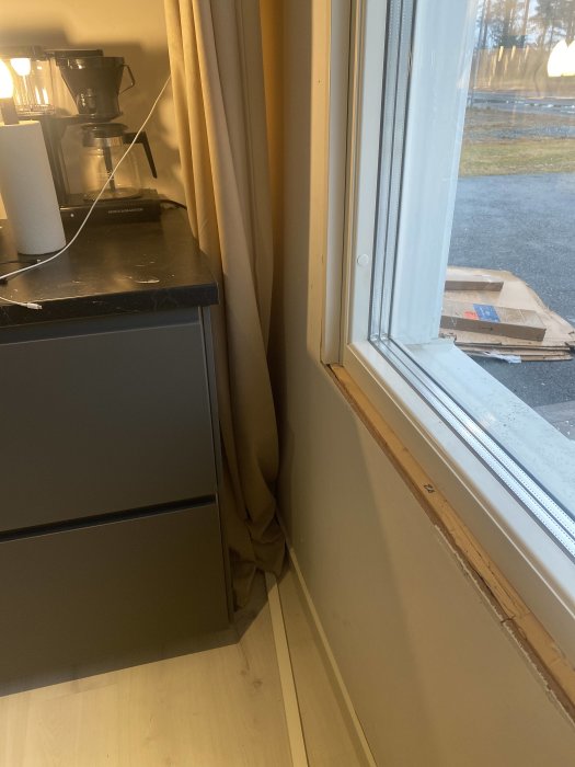 Inomhusvy: kaffebryggare på skåp, gardiner, fönster med utsikt, kartong på marken utanför.