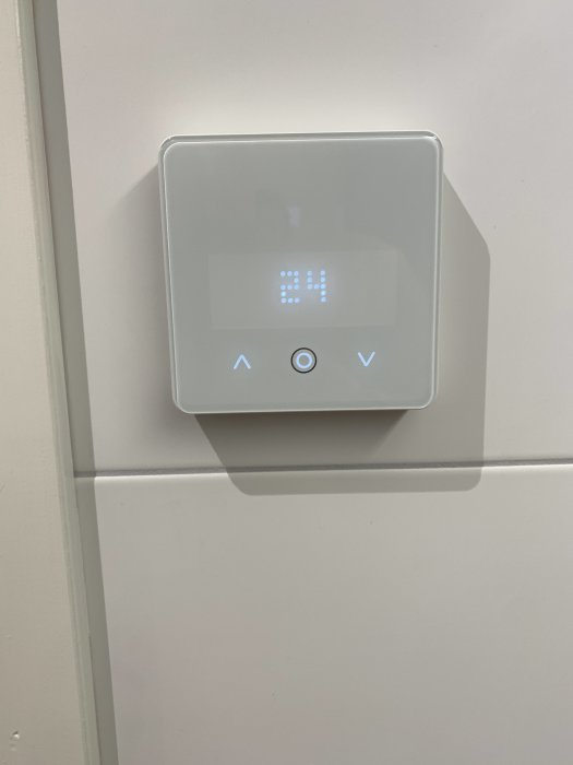 Vit termostat på vägg, digital display visar 24, upp/ner pilar, modern design.