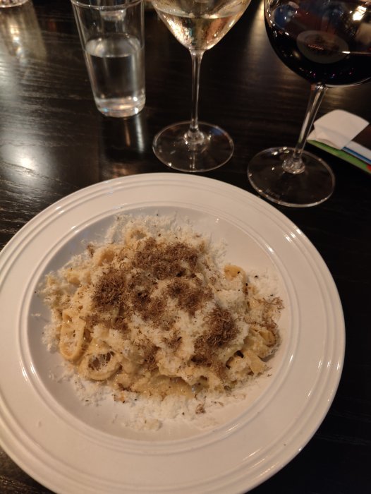 Pasta med riven ost och peppar, vin i glas, vattenglas, mörkt bord.