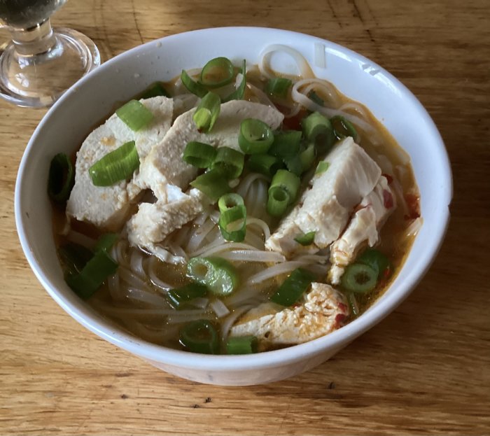 En skål med asiatisk nudelsoppa, kyckling, gröna lökar på träbord. Spicy och inbjudande.