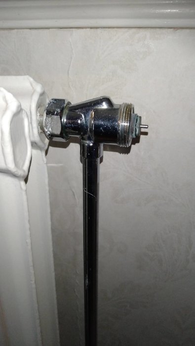 Termostatventil på radiator, vitmålad vägg, sprucken tapet, metall och svart rör.