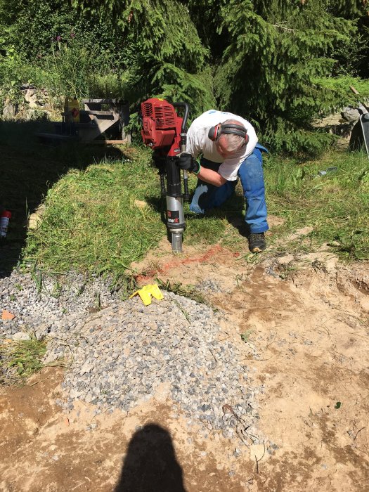 En person använder en motorborr för arbete utomhus. Skyddsutrustning, gräs, grus och trädfön bakgrund syns.