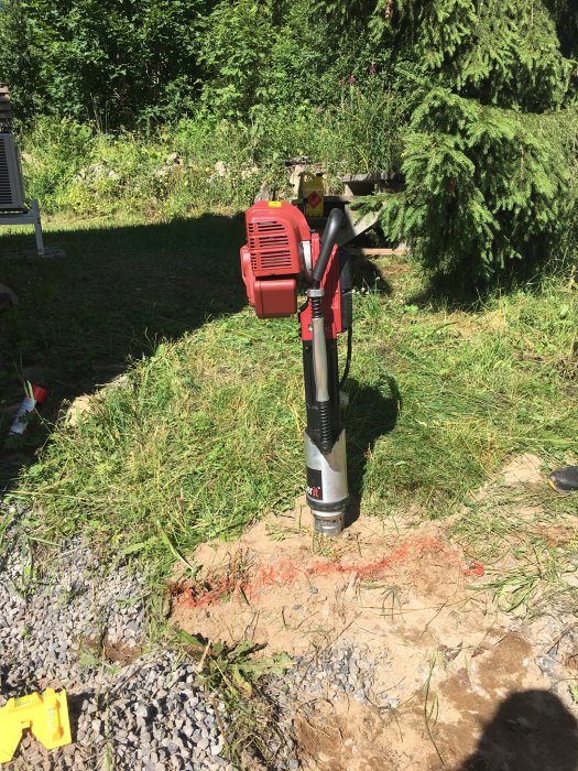 Motoriserad jordborr i gräsmiljö, grus och jord, verktyg synligt, träd och buskar i bakgrunden, soligt väder.