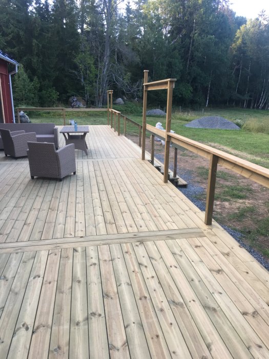 Trädäck med utemöbler vid hus, naturen i bakgrunden, oavslutad räckekonstruktion, dagtid.