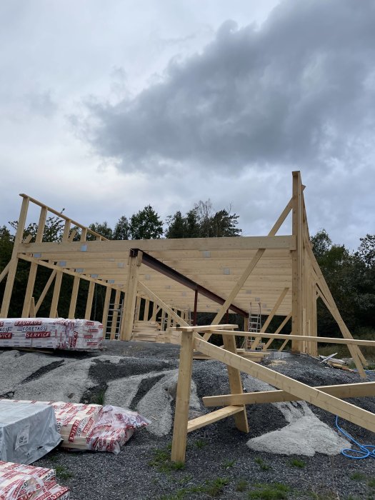 Byggplats med trästomme till en byggnad, molnig himmel, byggmaterial och grus på marken.